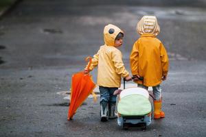 Two kids with umbrella, suitcase and yellow waterproof cloaks and boots walking outdoors after the rain together photo