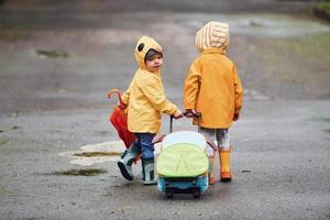 Two kids with umbrella, suitcase and yellow waterproof cloaks and boots walking outdoors after the rain together photo