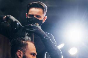 joven barbudo sentado y cortándose el pelo en la barbería por un tipo con máscara protectora negra foto