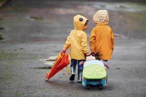 Two kids with umbrella, suitcase and yellow waterproof cloaks and boots walking outdoors after the rain together photo