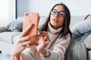 Making selfie. Young beautiful woman in glasses sitting at home alone with phone in hands photo