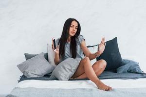 Holding phone and listens to music in wireless headphones. Young beautiful woman in casual clothes sitting at home alone on the bed photo