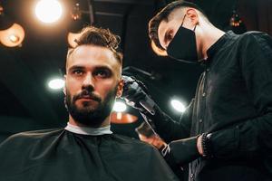 Front view of young bearded man that sitting and getting haircut in barber shop by guy in black protective mask photo