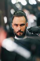 Young man with stylish hairstyle sitting and getting his beard shaved in barber shop photo