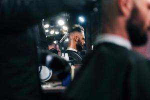 Young bearded man sitting and getting haircut in barber shop photo