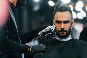 Young man with stylish hairstyle sitting and getting his beard shaved in barber shop photo