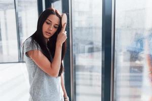 Beautiful young brunette in grey shirt standing near window and posing for a camera photo