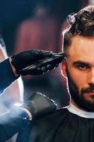 Close up view of young bearded man that sitting and getting haircut in barber shop photo