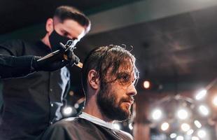 joven barbudo sentado y cortándose el pelo en la barbería por un tipo con máscara protectora negra foto