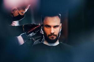 Front view of young bearded man that sitting and getting haircut in barber shop by guy in black protective mask photo