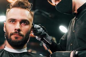 Front view of young bearded man that sitting and getting haircut in barber shop by guy in black protective mask photo