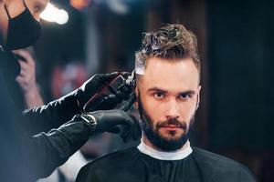 Front view of young bearded man that sitting and getting haircut in barber shop by guy in black protective mask photo