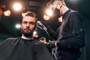 Front view of young bearded man that sitting and getting haircut in barber shop by guy in black protective mask photo