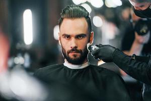 Young man with stylish hairstyle sitting and getting his beard shaved in barber shop photo