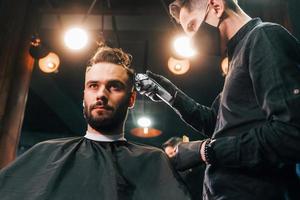 Front view of young bearded man that sitting and getting haircut in barber shop by guy in black protective mask photo