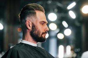 Young bearded man sitting in barber shop and waiting for stylist photo