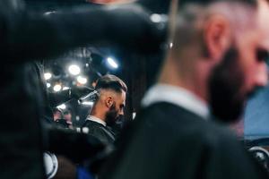 Young bearded man sitting and getting haircut in barber shop photo