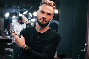 Young bearded man standing in barber shop and holding scissors photo