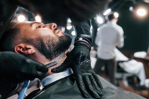 Young man with stylish hairstyle sitting and getting his beard shaved in barber shop photo