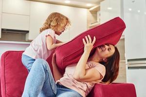 Having fun and jumping. Young mother with her little daughter in casual clothes together indoors at home photo
