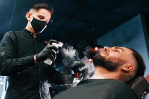 Young man with stylish hairstyle sitting and getting his beard shaved by guy in black protective mask in barber shop photo