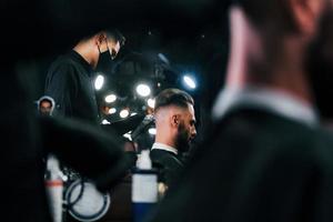 Young bearded man sitting and getting haircut in barber shop by guy in black protective mask photo