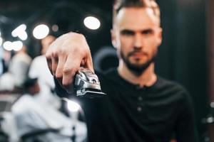 Young bearded man standing in barber shop and holding clipper photo