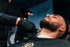 Young man with stylish hairstyle sitting and getting his beard shaved in barber shop photo