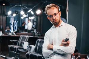 retrato de un joven vestido de blanco que está parado adentro en una barbería y sosteniendo un cortapelos foto