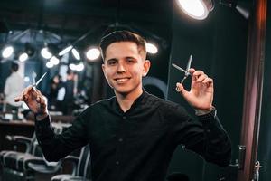 Portrait of young guy in black clothes that standing indoors in barber shop and holding scissors photo