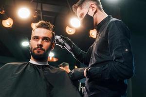 Front view of young bearded man that sitting and getting haircut in barber shop by guy in black protective mask photo