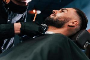 Young man with stylish hairstyle sitting and getting his beard shaved by guy in black protective mask in barber shop photo