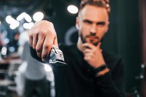 Young bearded man standing in barber shop and holding clipper photo