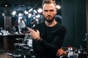 Young bearded man standing in barber shop and holding scissors photo