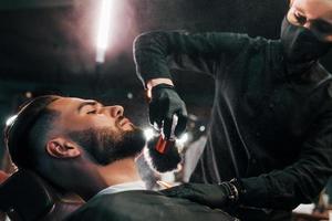 Young man with stylish hairstyle sitting and getting his beard shaved by guy in black protective mask in barber shop photo