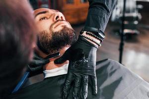 Young man with stylish hairstyle sitting and getting his beard shaved in barber shop photo