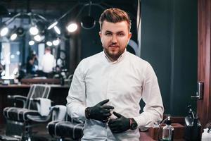 Portrait of young guy in white clothes and black gloves that standing indoors in barber shop and holding scissors photo