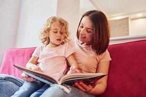 leyendo un libro grande. joven madre con su pequeña hija vestida de forma informal juntas en casa foto