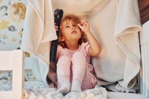 Sitting under blanket with flashlight. Cute little girl in casual clothes is indoors at home at daytime photo