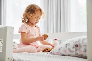 Sitting on the bed, eating cookies and drinking milk. Cute little girl in casual clothes is indoors at home at daytime photo