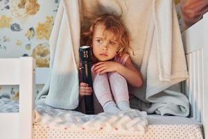 sentado debajo de una manta con una linterna. una niña linda con ropa informal está en el interior de su casa durante el día foto