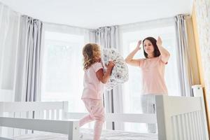 Having fun by throwing a pillow. Young mother with her little daughter in casual clothes together indoors at home photo