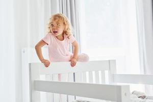 Standing on the bed. Cute little girl in casual clothes is indoors at home at daytime photo