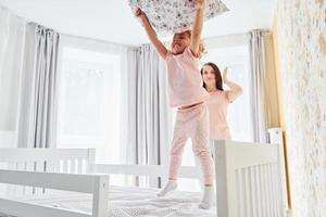 Having fun by throwing a pillow. Young mother with her little daughter in casual clothes together indoors at home photo