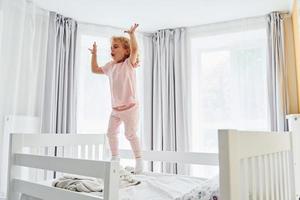 Jumping on the bed. Cute little girl in casual clothes is indoors at home at daytime photo