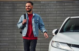 Handsome young guy in casual clothes is outdoors at sunny daytime near white car photo