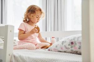 Sitting on the bed, eating cookies and drinking milk. Cute little girl in casual clothes is indoors at home at daytime photo