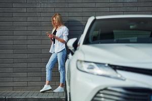 Beautiful blonde in casual clothes with phone near white car is outdoors at sunny daytime photo