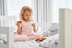 Having a breakfast by eating cookies. Cute little girl in casual clothes is indoors at home at daytime photo