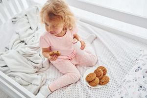 Having a breakfast by eating cookies. Cute little girl in casual clothes is indoors at home at daytime photo